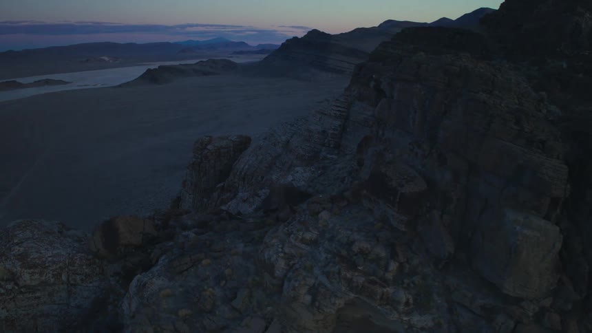 A slow, cinematic aerial shot pulls back from a rugged mountain cliff to reveal a vast, empty desert landscape at twilight