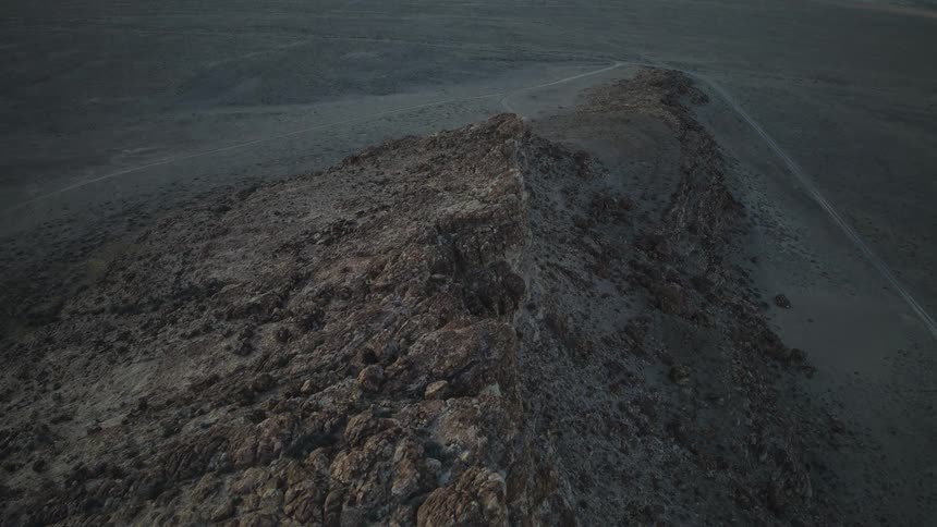 An aerial view slowly drifts along the top of a craggy rock ridge in a vast desert landscape at dusk