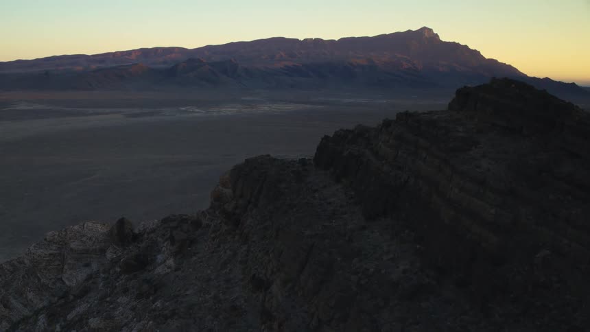 An aerial shot flies over a rocky mountain ridge overlooking a vast desert valley at sunrise