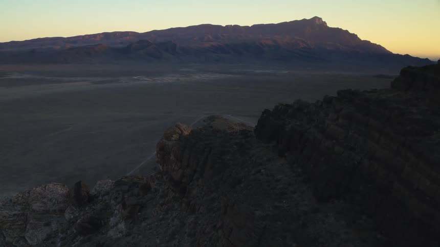 An aerial shot flies over a rocky mountain ridge overlooking a vast desert valley at sunrise