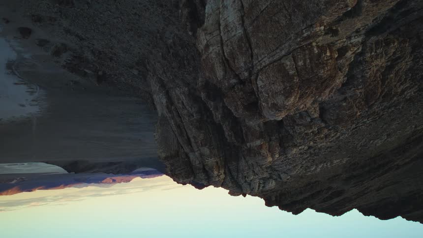 A vertical timelapse shows the shifting light of sunset over a vast desert landscape with a prominent rocky cliff in the foreground