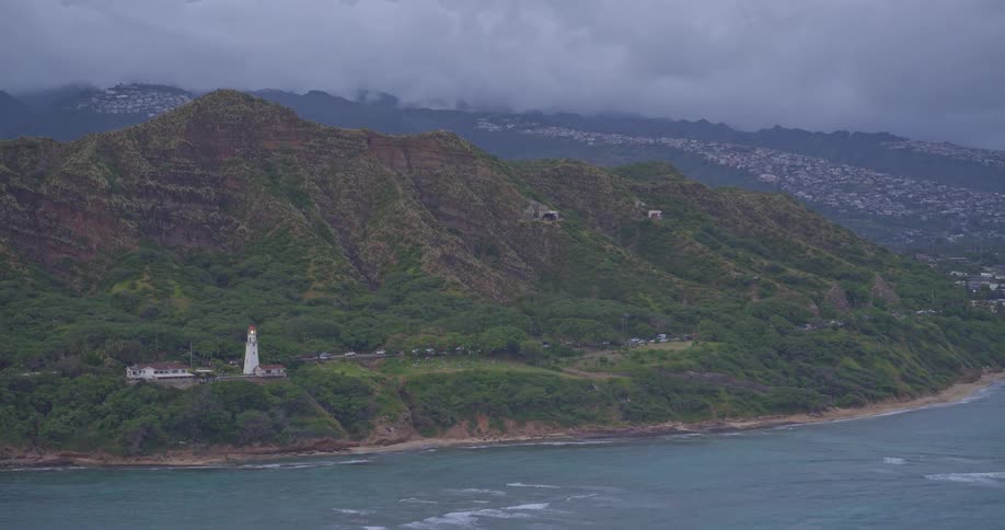A slow pan reveals the Diamond Head Lighthouse on the lush, green coast of Oahu, Hawaii, with its light flashing under a moody, overcast sky