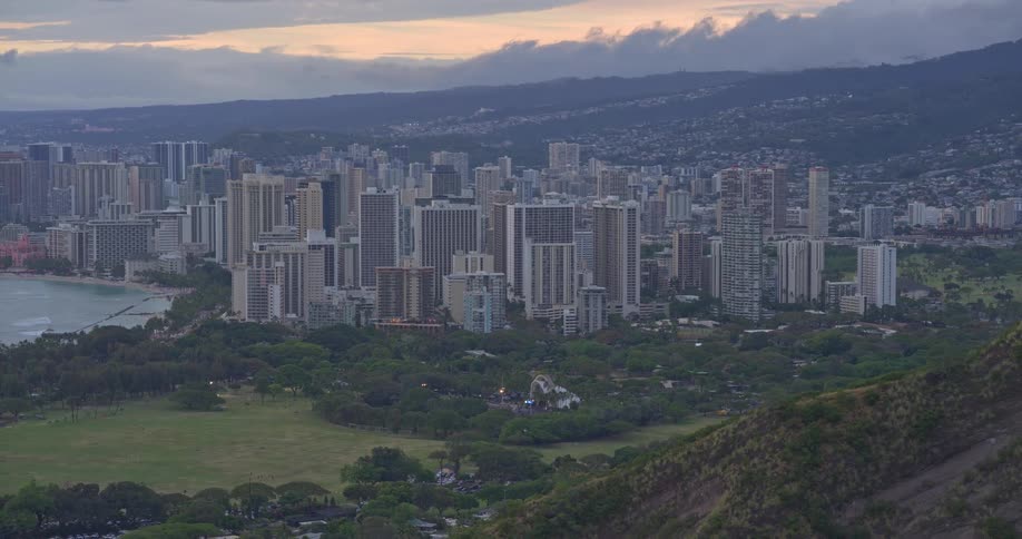 A sweeping scenic view of the Honolulu city skyline and Waikiki Beach from a high vantage point as evening sets in