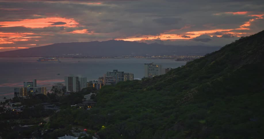 A slow pan reveals the glowing city of Honolulu next to the ocean, with the Diamond Head lighthouse in the foreground during a vibrant sunset