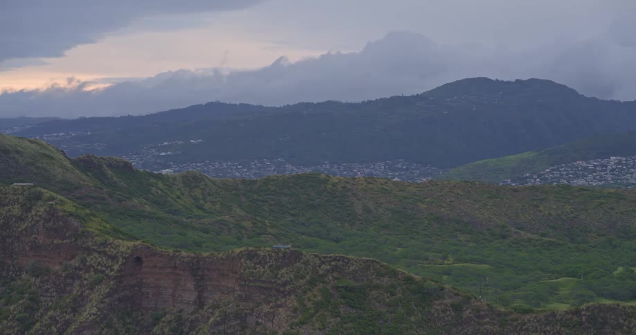 A slow zoom out reveals the vast, lush green mountains overlooking a distant city under a cloudy, overcast sky