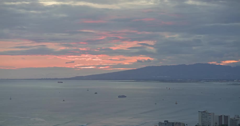 A serene, wide shot of a vast ocean with boats scattered on the surface under a cloudy sky painted with the pink hues of sunset