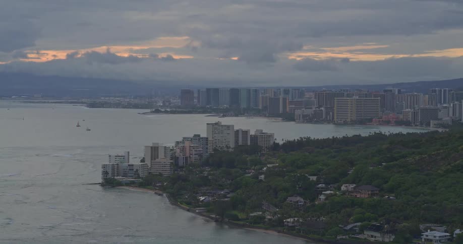A serene wide shot of the Honolulu, Hawaii coastline and cityscape under a moody, cloudy sky at sunset