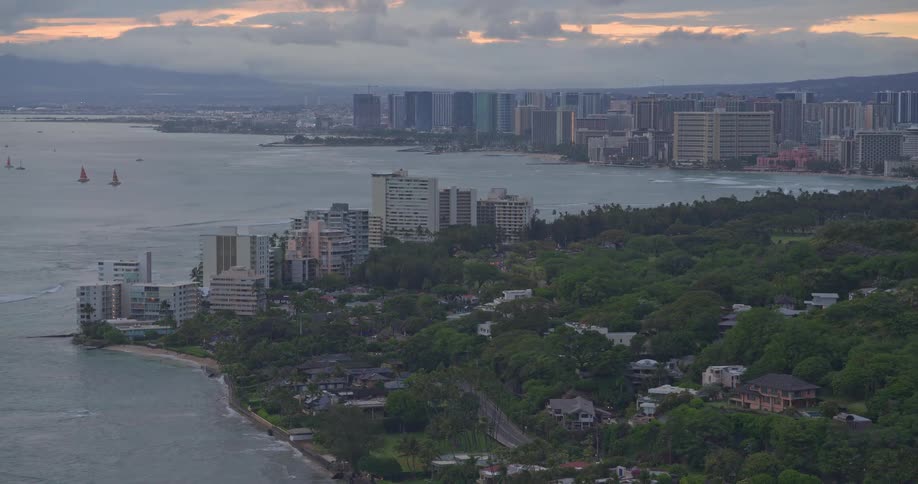 An elevated view of the Honolulu, Hawaii coastline shows the city skyline and lush greenery under a cloudy sky at dusk