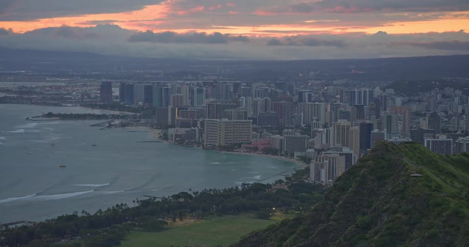 A stunning pull-out shot from the top of Diamond Head reveals the Honolulu city skyline and Waikiki beach under a beautiful cloudy sunset