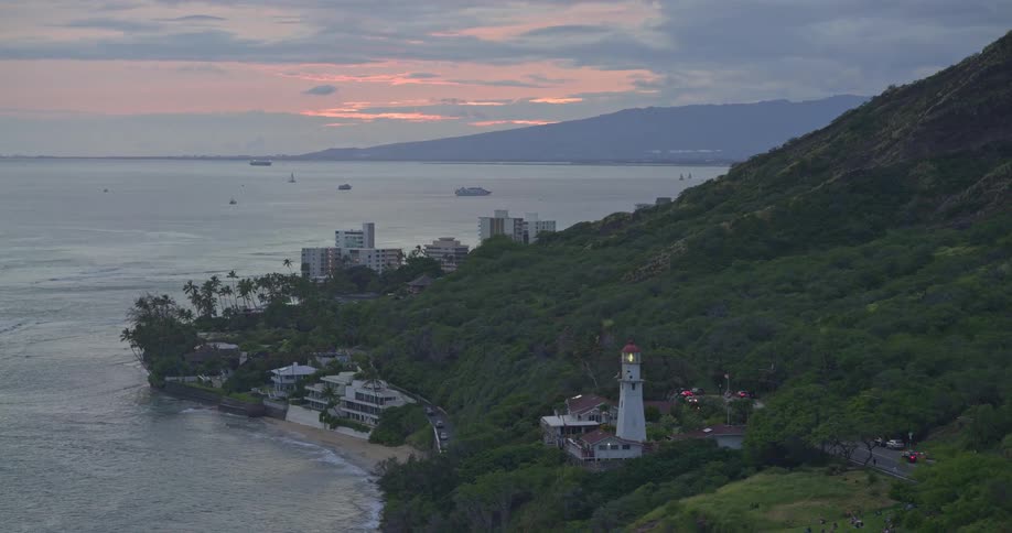 The Diamond Head Lighthouse illuminates the lush Hawaiian coastline at sunset as boats sail on the Pacific Ocean