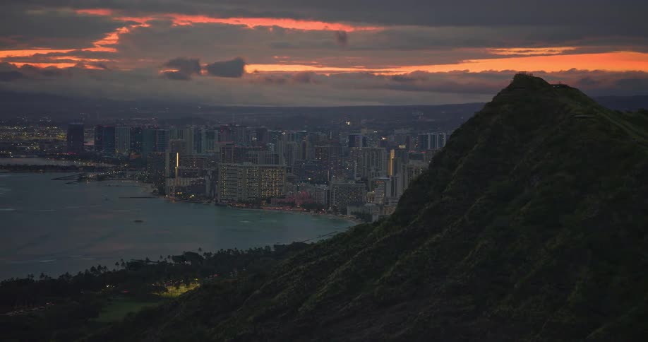 A breathtaking aerial view from a lush mountain reveals the Honolulu city skyline along the coast at twilight under a dramatic, colorful sky