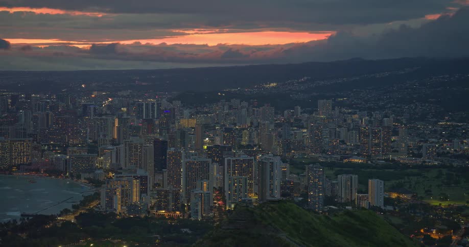 A stunning panoramic view of the illuminated Honolulu city skyline and Waikiki Beach at dusk under a beautiful orange sunset