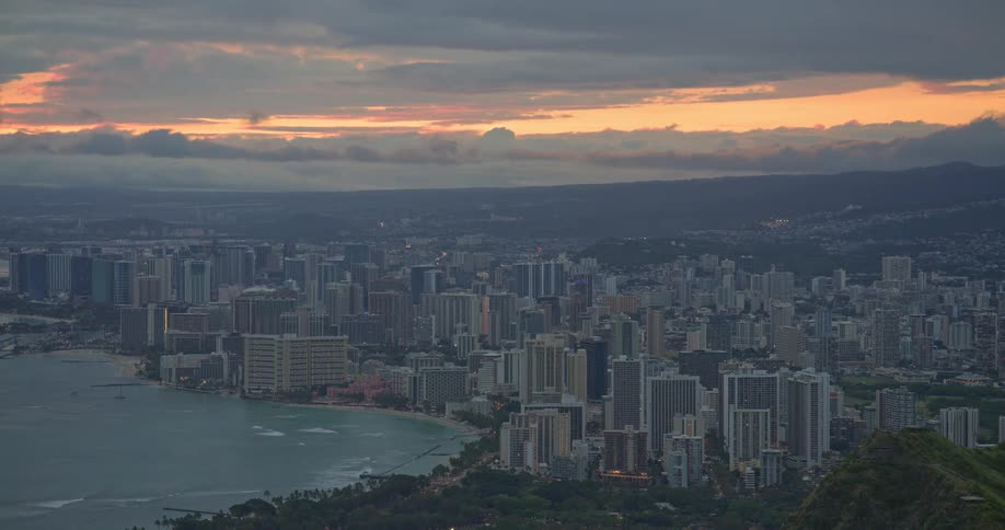 A high-angle, pull-back shot reveals the city of Honolulu along the Waikiki coastline under a dramatic, cloudy sunset sky in Hawaii