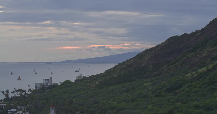 A serene view of the Hawaiian coast as sailboats dot the ocean next to a lush mountain at dusk