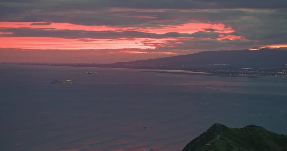 A cargo ship sails across the calm ocean as a vibrant red sunset lights up the cloudy sky over a distant coastline