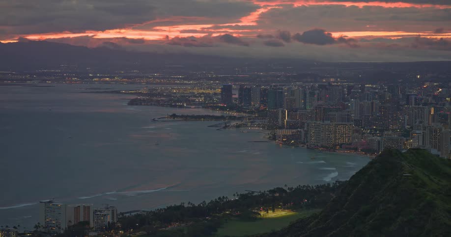 The city lights of Waikiki and Honolulu glow along the coastline at dawn, viewed from a high vantage point during a dramatic sunrise