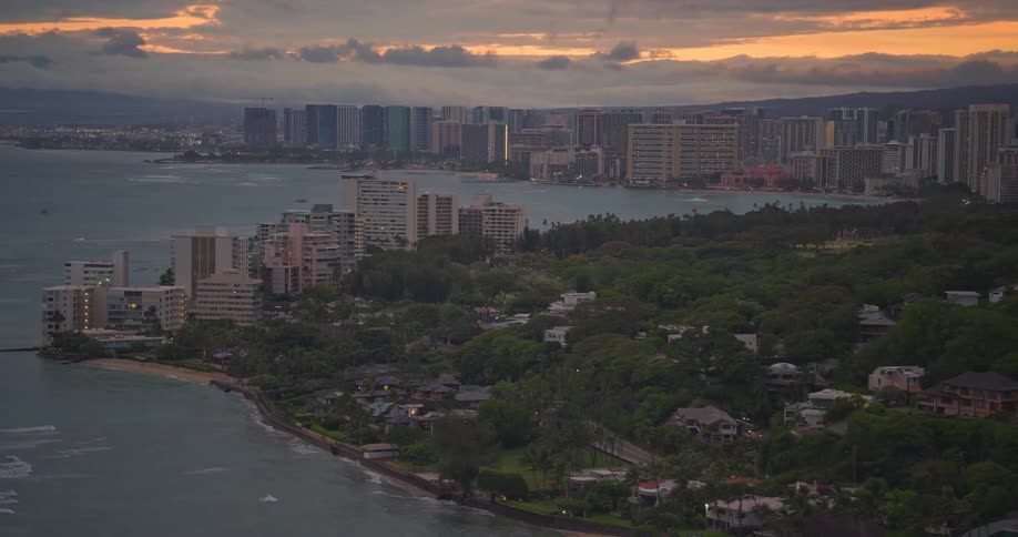 A scenic aerial pan across the Honolulu and Waikiki coastline reveals the city skyline against a dramatic sunset