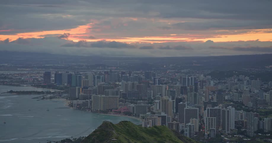A panoramic high-angle view of the city of Honolulu and Waikiki Beach at sunset with a warm glow on the horizon