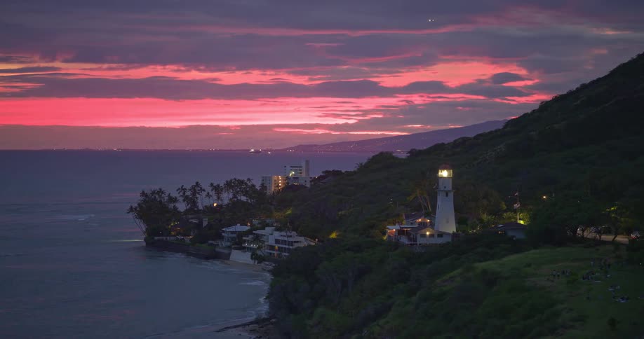 A beautiful lighthouse on a lush green cliffside illuminates the coastline under a vibrant pink and purple sunset sky