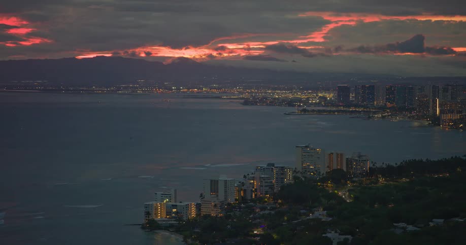 A high-angle view of a coastal city illuminated at night, with a fiery red sunset glowing on the horizon over the ocean