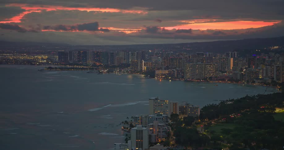 An aerial view of the illuminated Waikiki skyline along the Honolulu coast at dusk under a colorful sunset sky