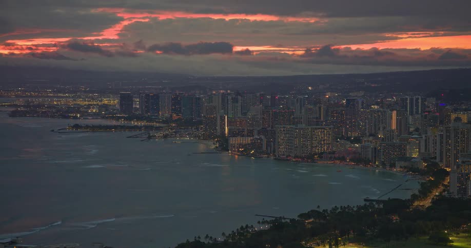 An expansive aerial view captures the illuminated Waikiki, Hawaii cityscape and coastline at dusk under a fiery sunset