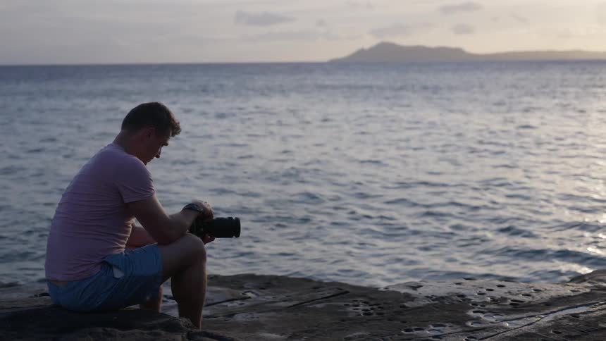 A solitary photographer sits on a rocky shoreline, adjusting his camera settings as he gazes at the tranquil ocean during sunset