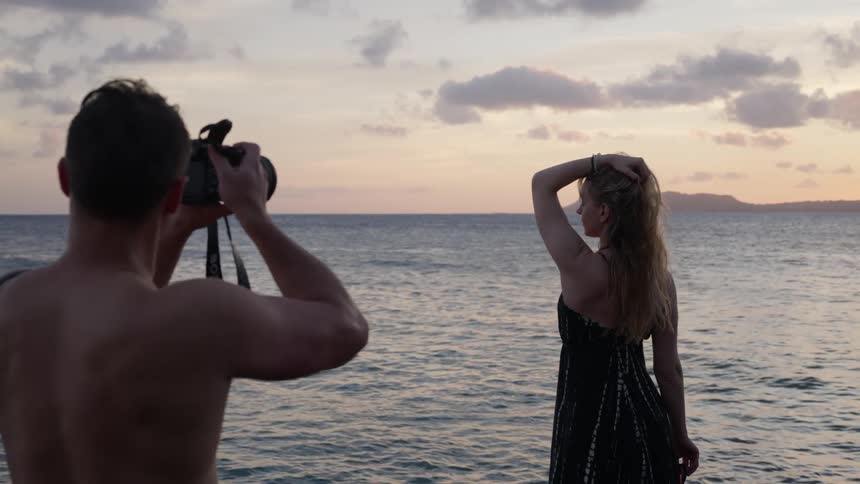 A shirtless photographer captures images of a woman posing in a dress against the backdrop of a serene ocean sunset