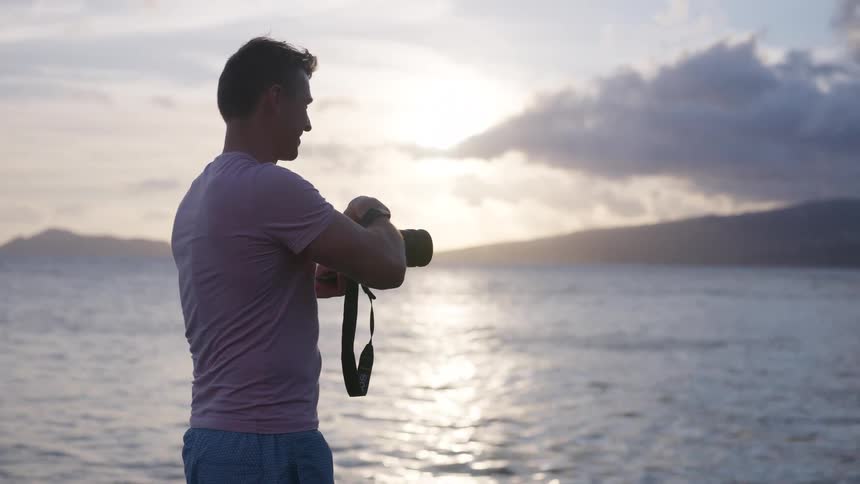 A photographer checks his camera settings while standing on the coast, backlit by the setting sun over the ocean
