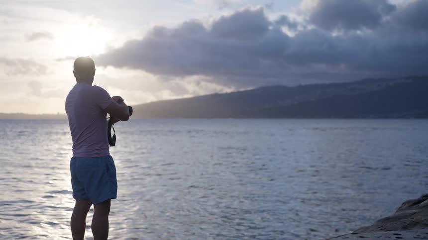 A male photographer stands silhouetted on a rocky shore, adjusting his camera settings while overlooking the ocean at sunset