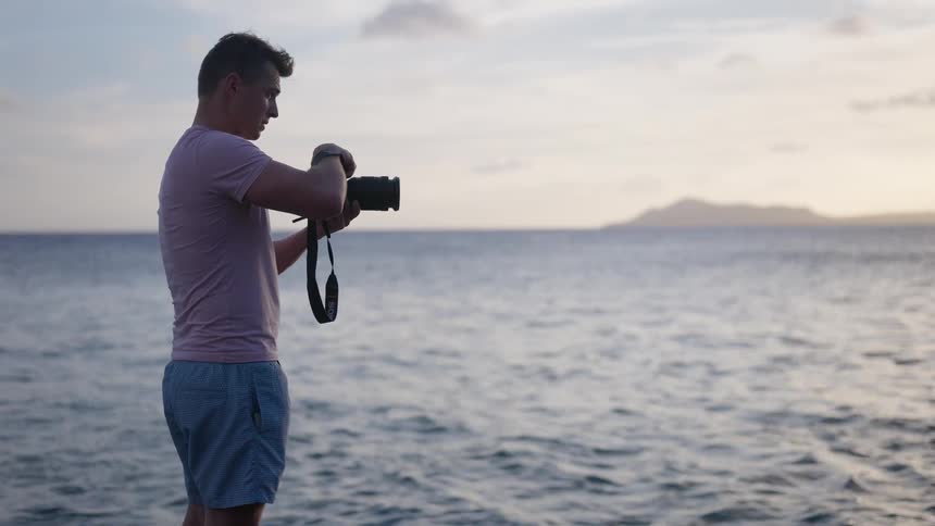 A young male photographer adjusts his camera settings while standing by the ocean at sunset