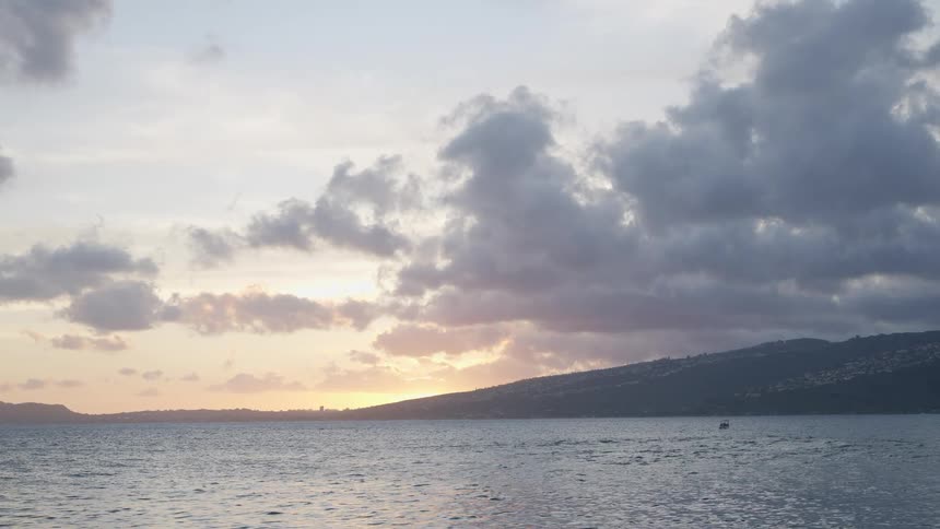 A calm ocean reflects the beautiful cloudy sky as the sun sets behind a distant, lush island