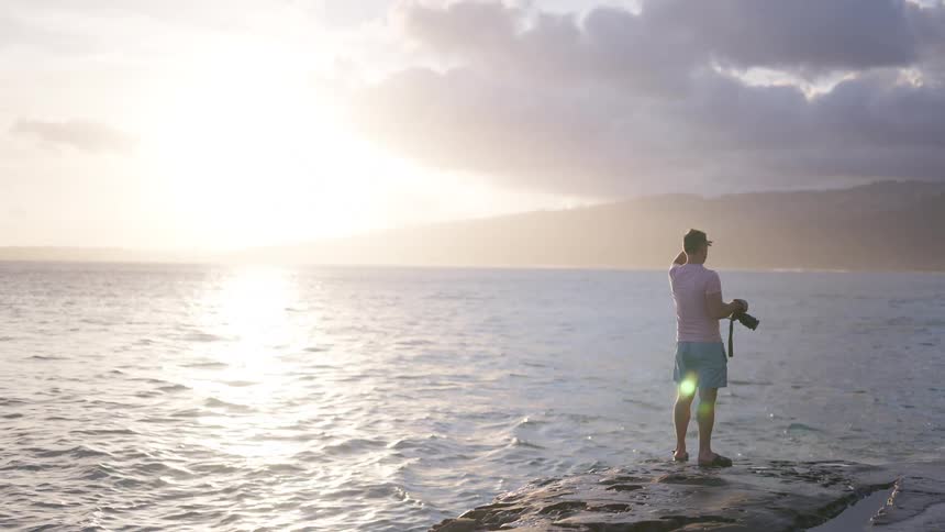 A male photographer stands on a rocky pier, looking out at the beautiful ocean view during a vibrant sunset