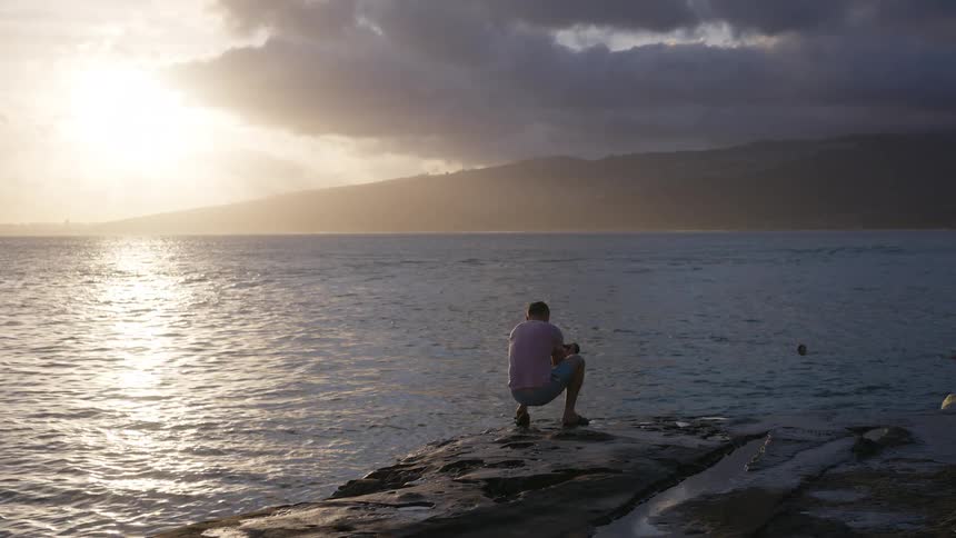 A solitary man squats on a rocky coast, peacefully watching the golden sunset reflect on the ocean