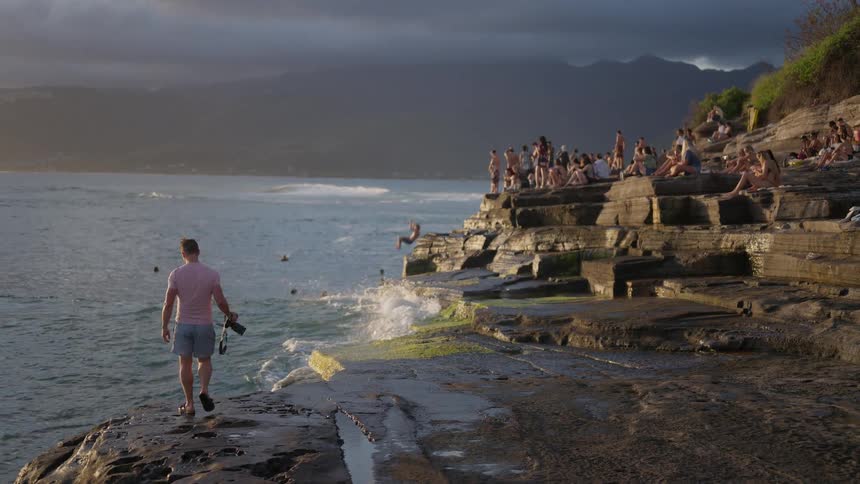 A photographer watches as a man cliff jumps into the ocean in slow motion while a crowd looks on from the rocky shoreline at sunset