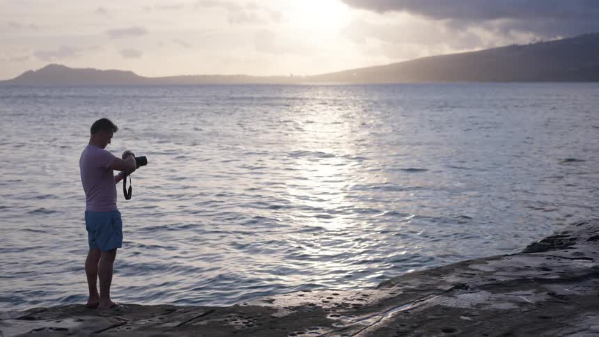 A male photographer stands on a rocky ledge, preparing his camera to shoot the stunning sunset over the water