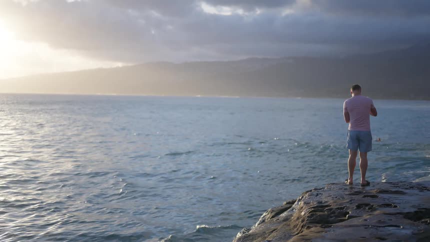 A man stands alone on a rocky point overlooking the vast ocean as waves splash at his feet during a hazy sunset