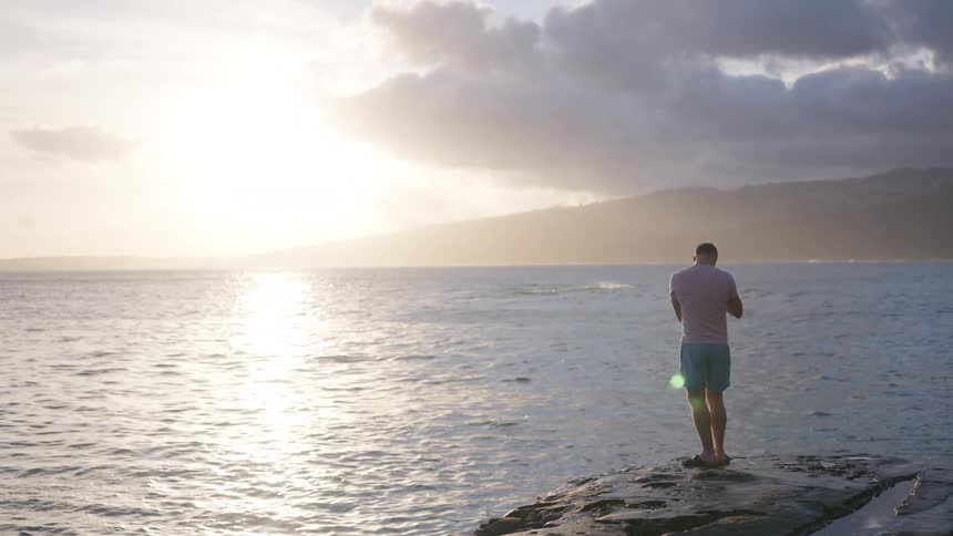 A male photographer stands on a rocky shore, adjusting his camera to capture the beautiful ocean landscape at sunset