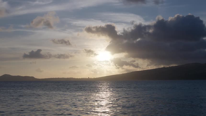 A serene sunset reflects on the calm ocean with silhouetted hills on the horizon under a cloudy sky