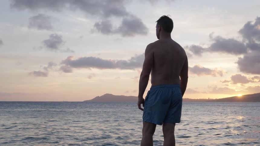 A man in swim trunks stands with his back to the camera, watching the serene sunset over the ocean