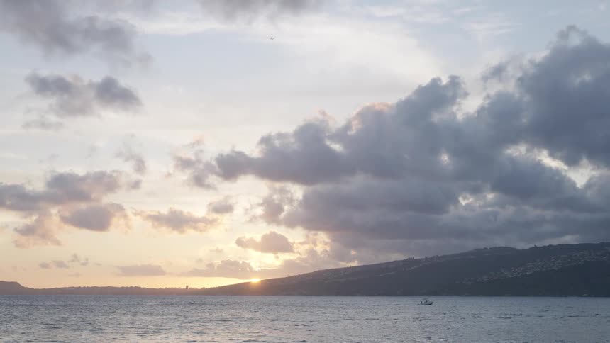 A small boat cruises across the tranquil ocean as the sun sets behind a cloud-covered island coastline