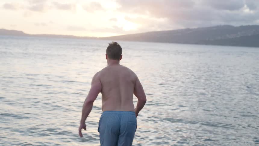 A shirtless man takes a running leap off a rocky pier and splashes into the ocean at sunset in slow motion