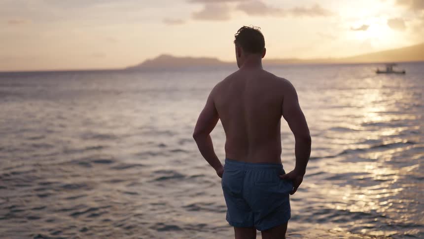 A shirtless man stands alone by the water's edge, looking out at the ocean during a golden sunset