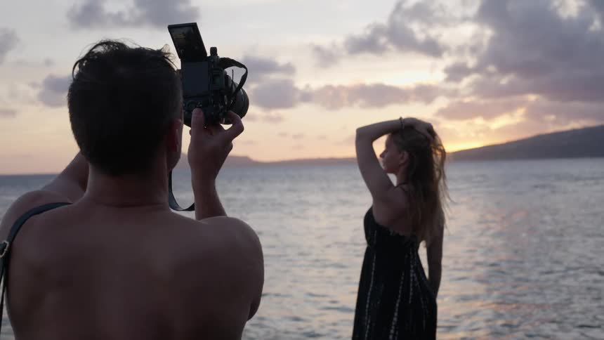 Over-the-shoulder slow motion shot of a shirtless photographer taking pictures of a woman posing by the ocean at sunset