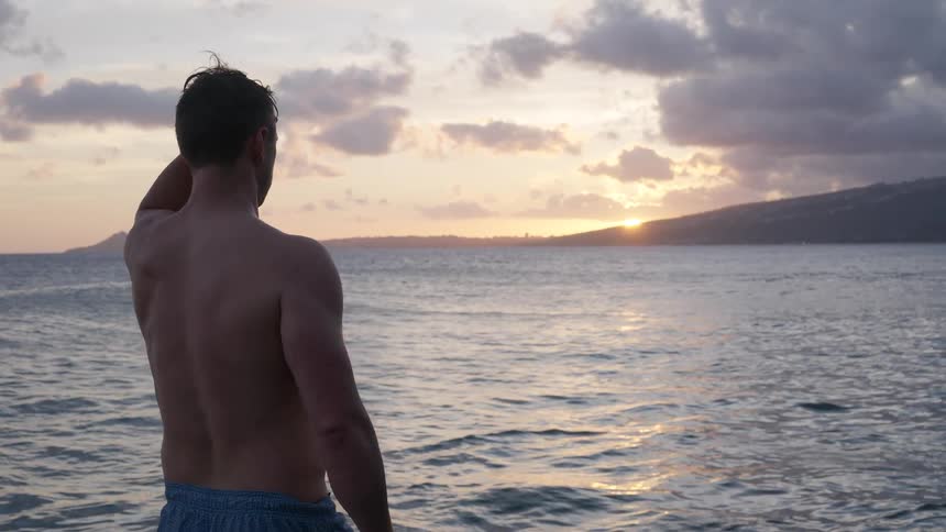 A contemplative shirtless man stands by the calm ocean watching the beautiful sunset over the horizon in slow motion