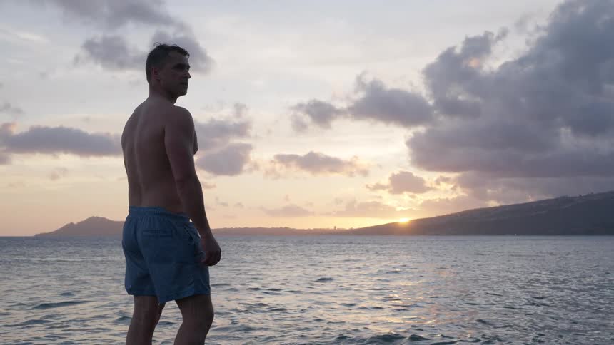 A lone man in swim trunks stands at the oceans edge, watching the beautiful sunset before turning and stretching in slow motion