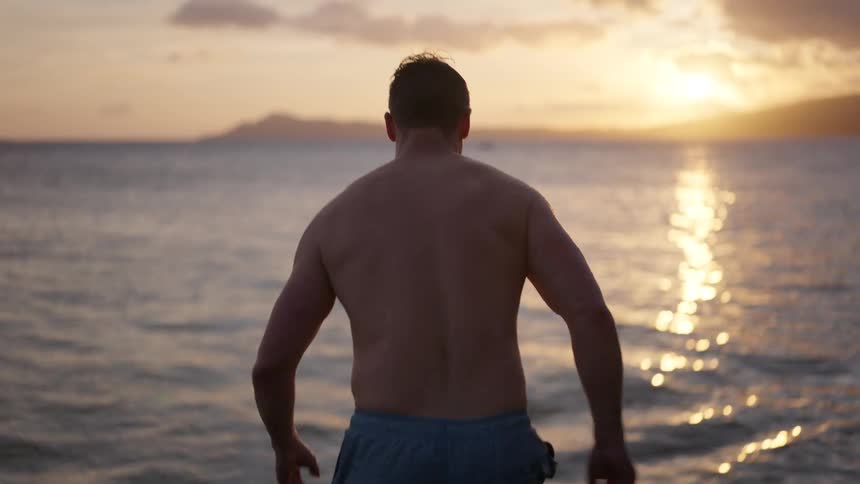 A man takes a dramatic running leap off a rocky ledge and into the ocean at sunset in slow motion