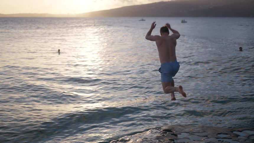 A man in swim trunks runs and jumps off a rocky ledge into the ocean at sunset in slow motion