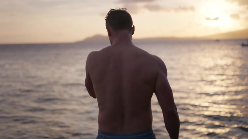 Slow motion rear view of a man in swim trunks looking out at the ocean during a golden sunset