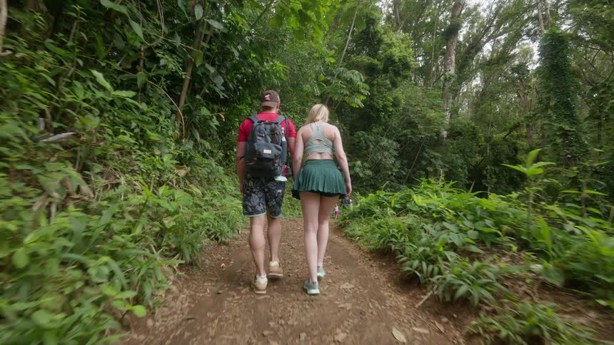 A young couple holds hands as they walk along a dirt path through a lush green jungle in slow motion